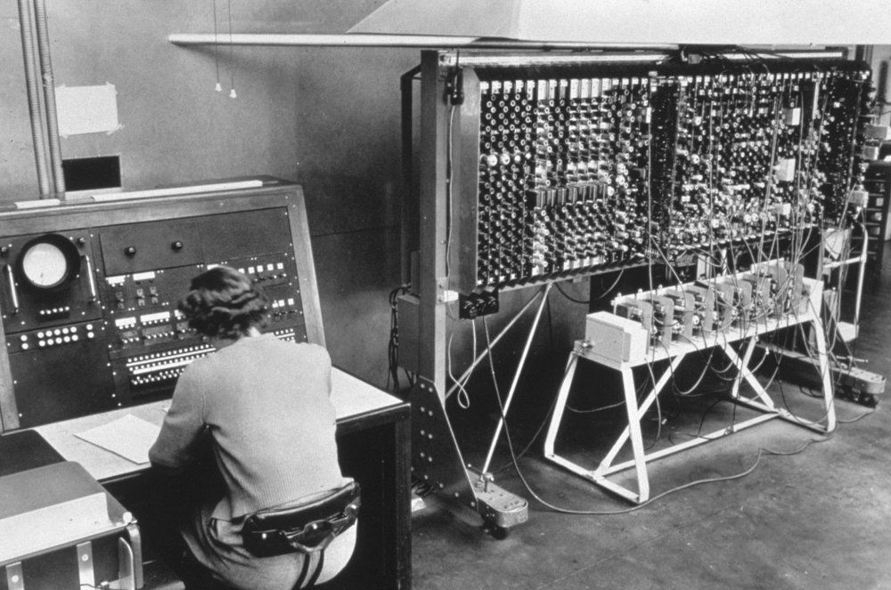 Black-and-white photograph of an operator seated at the control desk of an early electronic computer, beside a large rack of valves and patch wiring.
